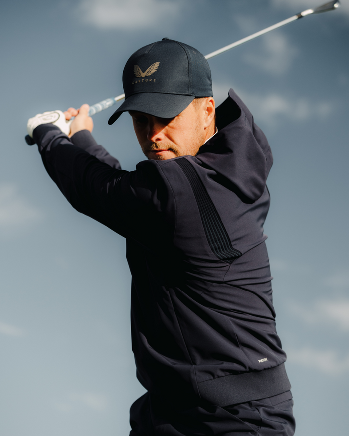 Male golfer in midnight navy performance hoodie mid swing against a cloudy sky
