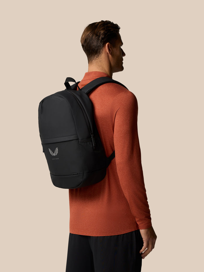 Back view of a man wearing a gunmetal Castore Core Backpack over an orange top.