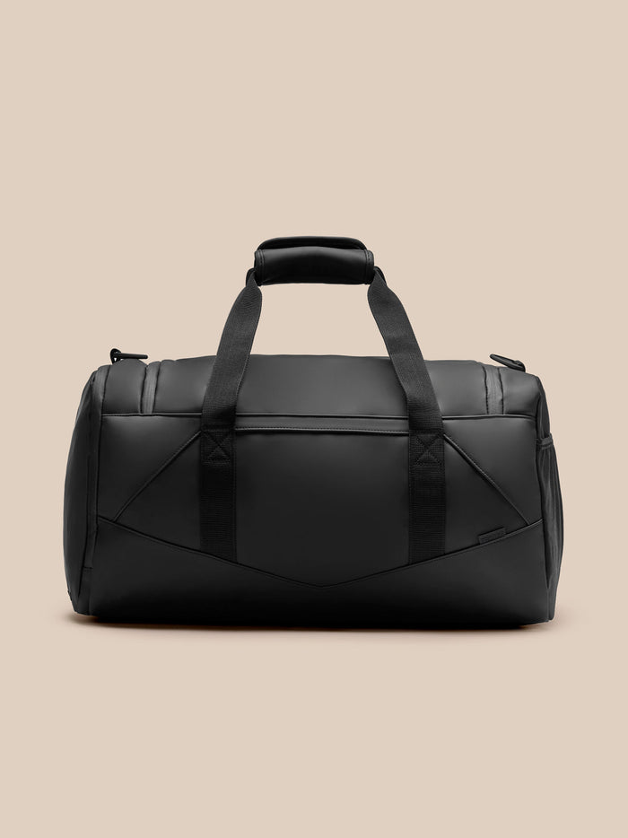 Core Holdall in black with padded top handle and sturdy webbing straps, front view against a beige background