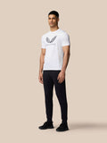 Male model wearing white Castore graphic training T shirt with wings logo on chest and black pants against a beige background.