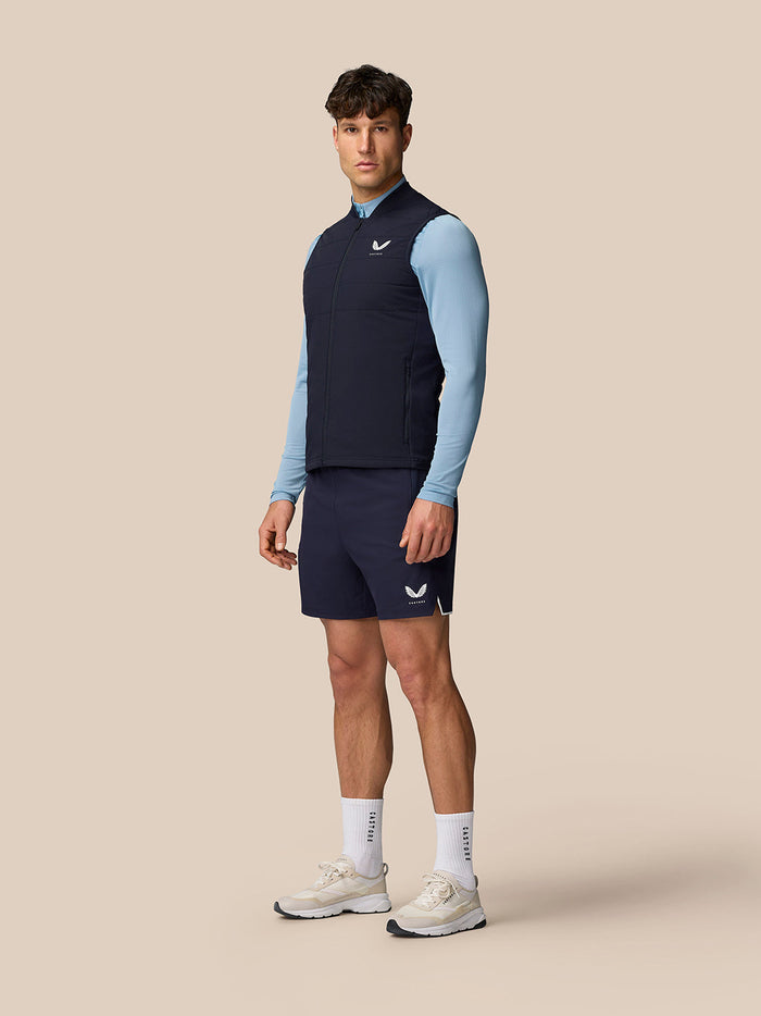 Model wearing navy Apex Training Gilet over a light blue long sleeve top and navy shorts with Castore logo on the chest