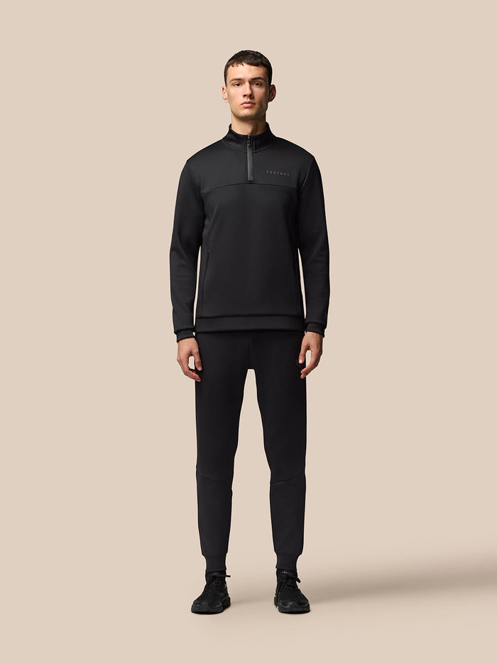 Full-body shot of male model in black Flex Lifestyle 1/4 Zip top with matching joggers against a beige backdrop.