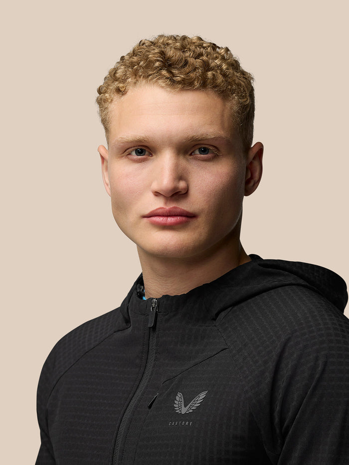 Front view of a model wearing a black Flex Winterised Jacket with hood, zipped up and chest logo visible.