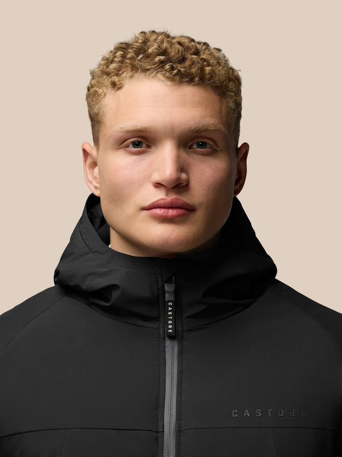Close-up of a man in a black longline wadded jacket with front zip and hood branding visible