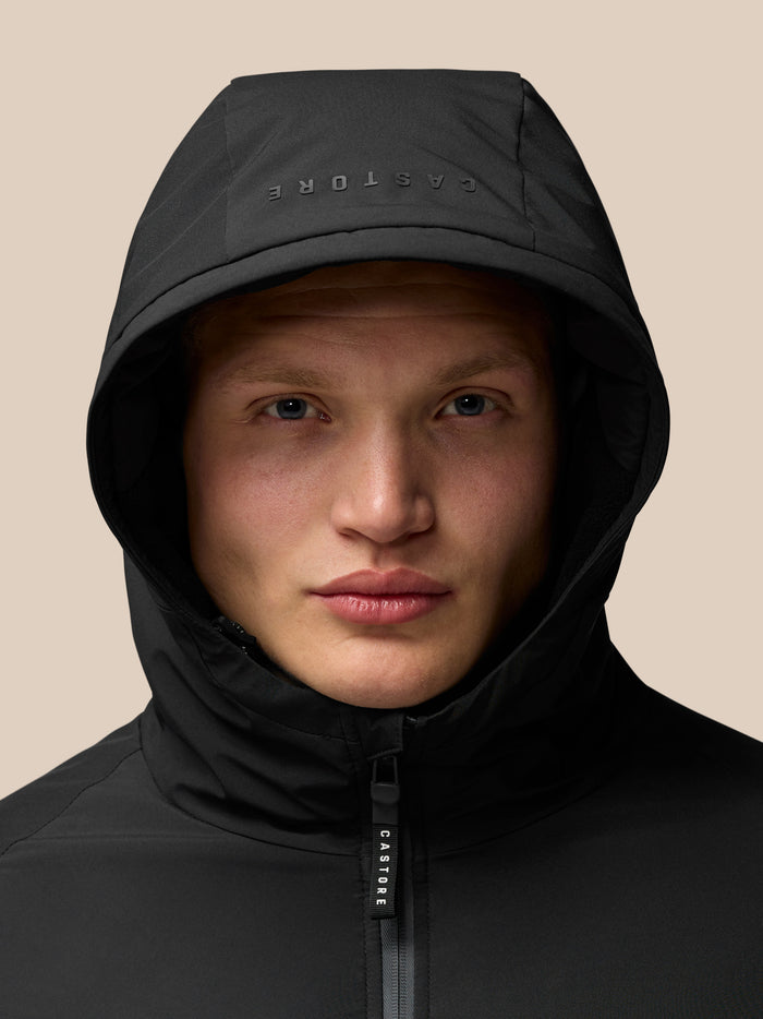 Model wearing a black hooded jacket with the hood up and a Castore zipper pull visible against a beige background
