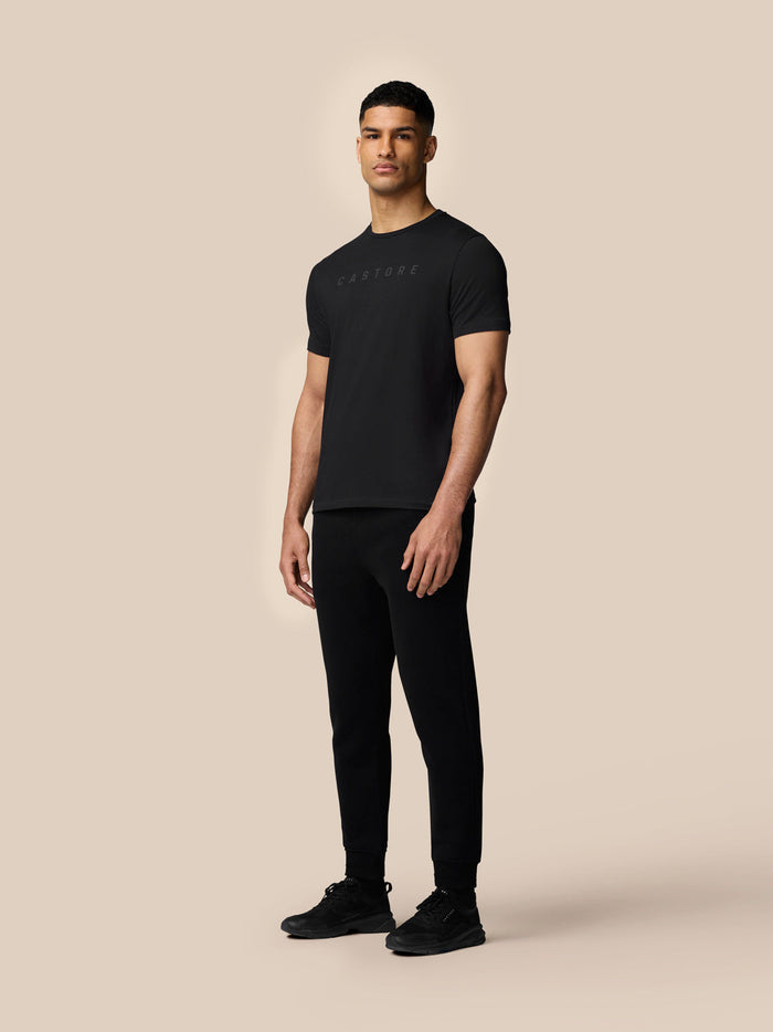 Male model in black Graphic Lifestyle T-Shirt with Castore graphic on the front against a beige backdrop.