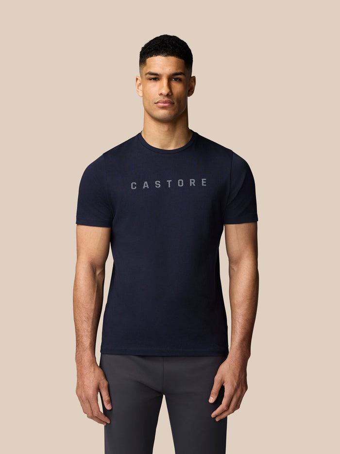 Male model wearing navy graphic Castore T shirt with crew neck and drop shoulders against a beige background