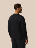 Three quarter rear view of a man in a black crew neck sweatshirt showing a small white wing logo near the hip