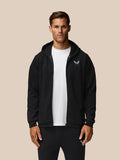 Male model wearing black Travel Lifestyle Zip Hoodie with white tee and Castore logo on the left chest against a beige background.