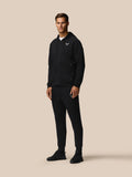 Full body shot of male model in black Travel Lifestyle Zip Hoodie and matching joggers with Castore logos on chest and leg against beige background.