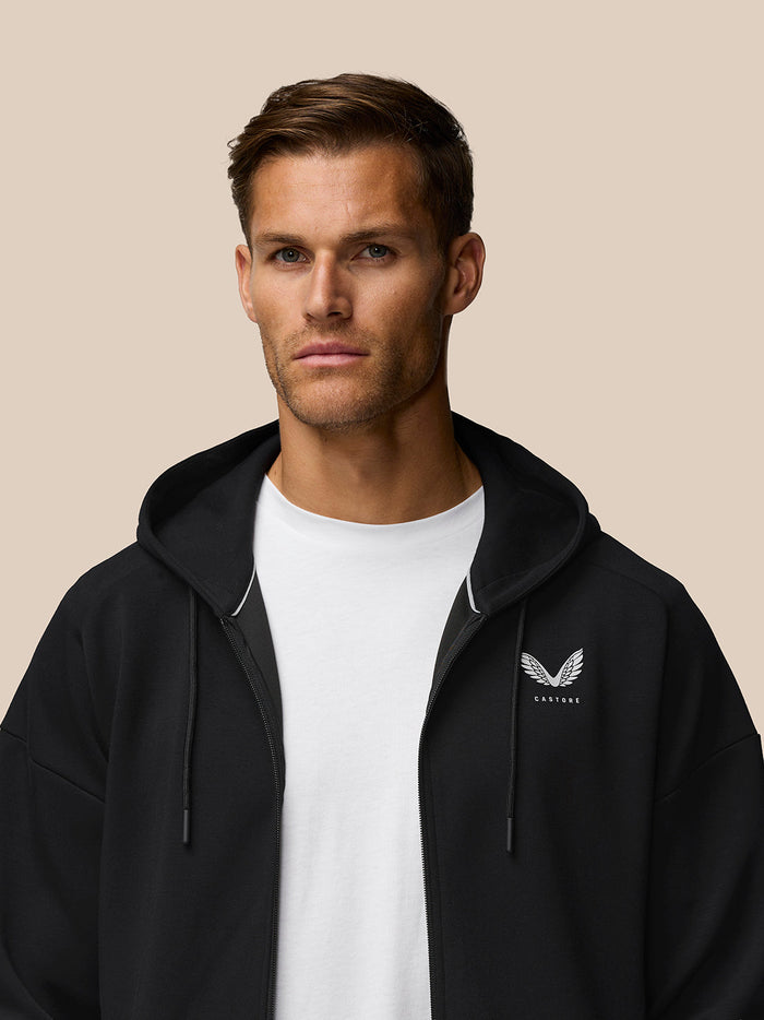 Close up of male model in black Travel Lifestyle Zip Hoodie with hood on and Castore logo on the chest.