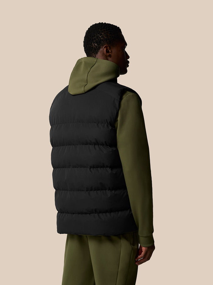 Back view of a man wearing a black sleeveless puffer gilet over an olive green hoodie.