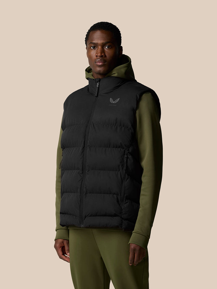 Front view of a man wearing a black puffer gilet over an olive green hoodie against a beige backdrop.
