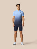 Male model wearing a light blue gradient polo and navy golf shorts with white shoes against a beige background.