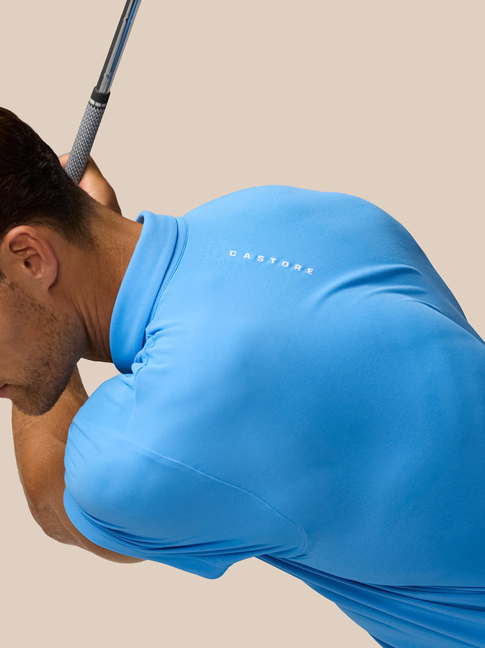 Male golfer wearing a blue Castore Golf Club Essential Polo with logo on chest mid backswing against a beige background