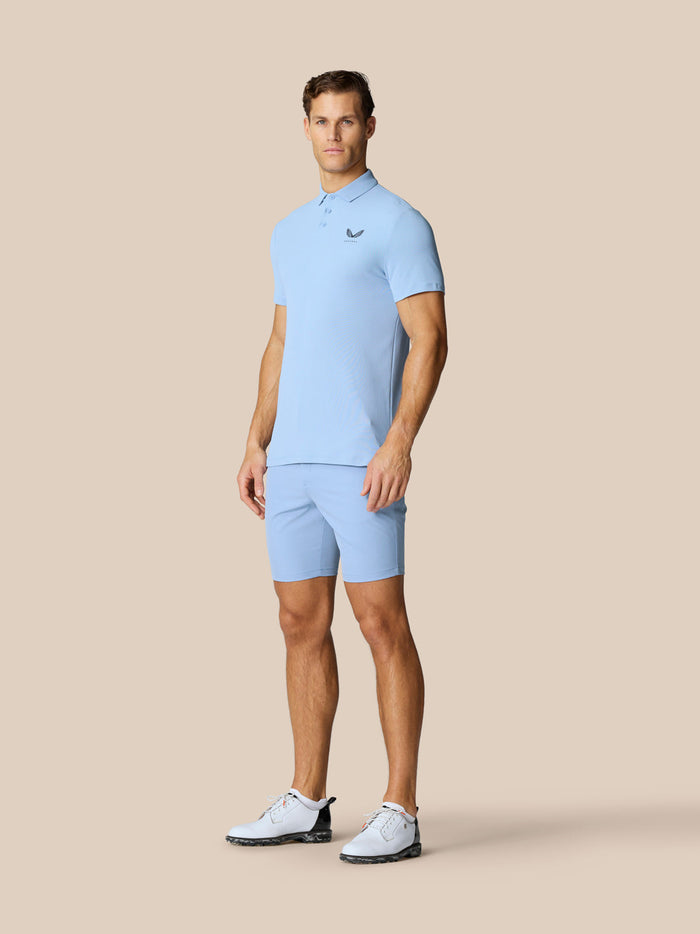 Male model in sky blue Golf Club Essential Polo with matching shorts and white golf shoes.