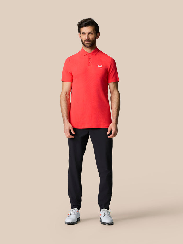 Full body shot of a man in a red Golf Club Essential Polo with dark trousers and white golf shoes.