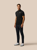 Full-body shot of a man in a black Pinnacle Pique Stripe Polo with Castore logo, paired with dark trousers and white golf shoes