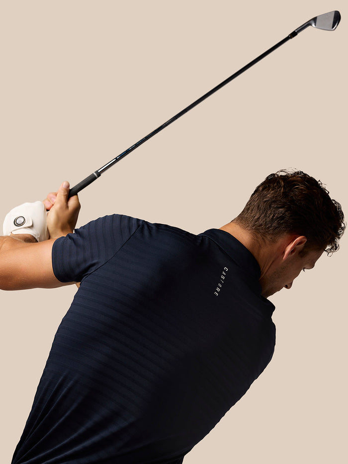 Golfer wearing a navy Pinnacle Pique Stripe Polo mid swing against a neutral beige background