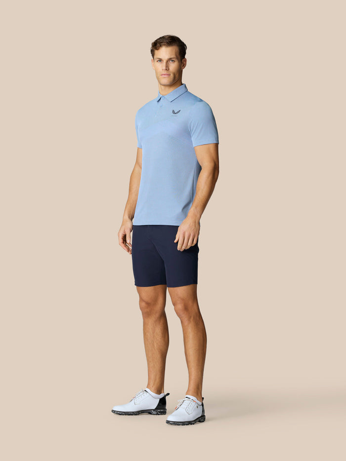 Man wearing sky blue Golf Pinnacle Polo with navy shorts on a beige background