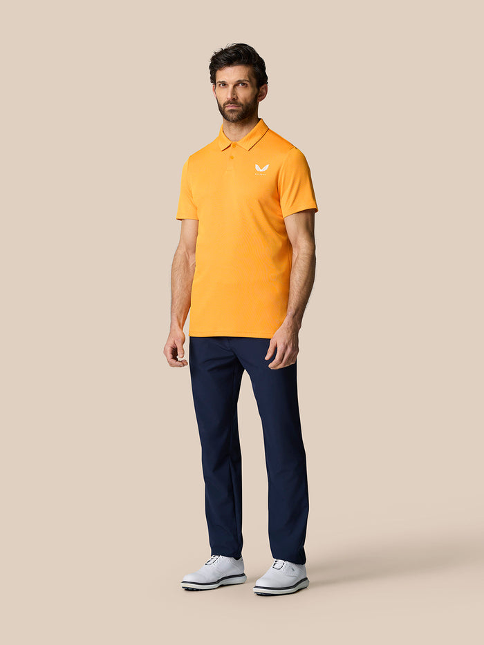 Full body shot of a man wearing the Golf Pinnacle Polo Mango in orange with navy trousers and white shoes.