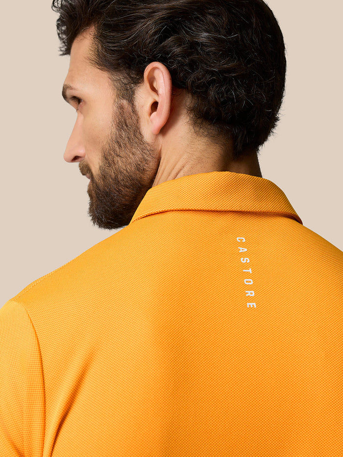 Back view of a model wearing Golf Pinnacle Polo Mango in orange with Castore branding on the upper back.
