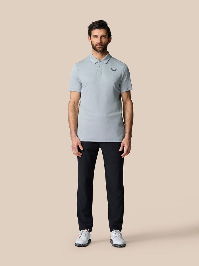 Full body shot of a man in a mid grey golf polo with a chest logo, dark trousers and white golf shoes.