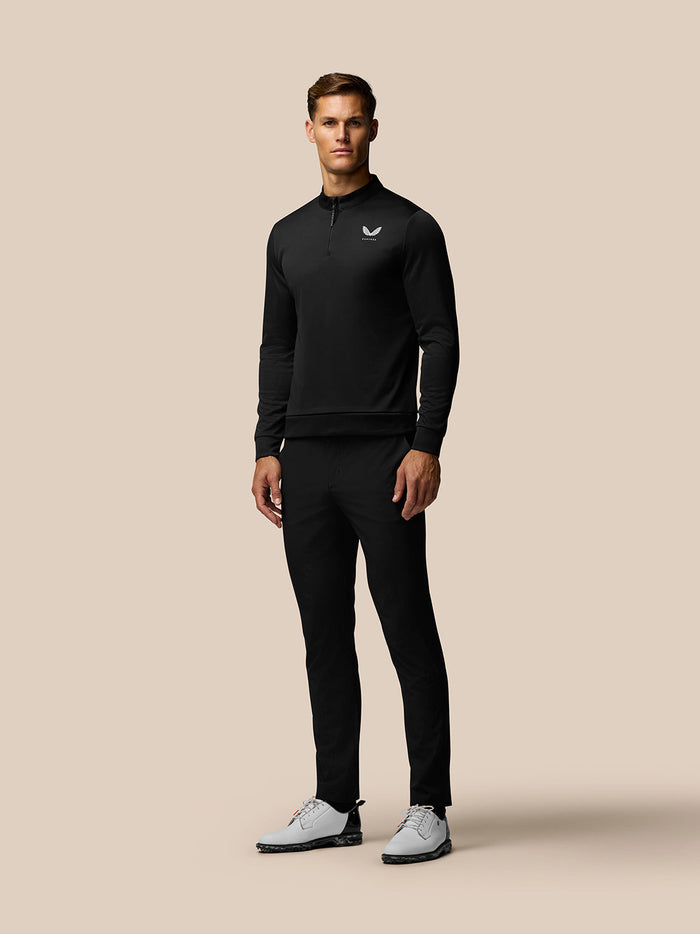 Male model wearing black Golf Club Classic 1/4 Zip Top with stand collar and Castore logo on chest, paired with white golf shoes.