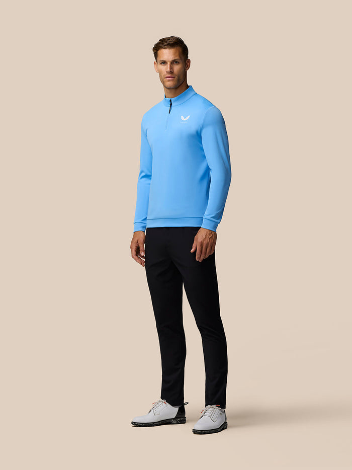 Full-length shot of a man in a light blue Golf Club Classic quarter zip top with front zipper left chest logo and white cap against a beige backdrop