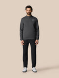 Full-body view of a man wearing a dark gray Castore golf quarter zip pullover with a chest logo, black pants and white golf shoes.