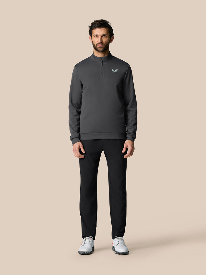 Full-body view of a man wearing a dark gray Castore golf quarter zip pullover with a chest logo, black pants and white golf shoes.