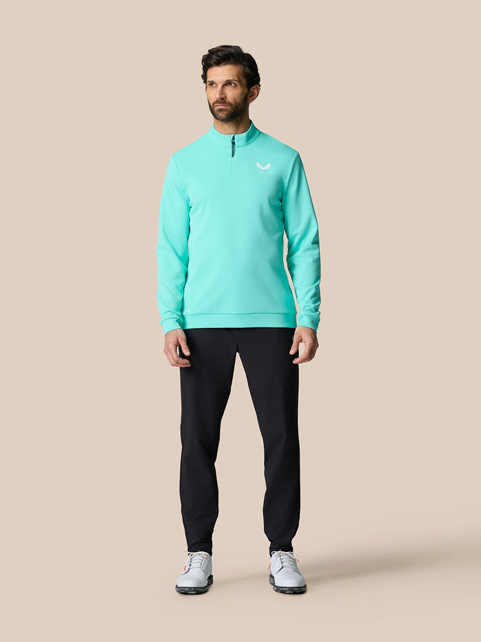 Male model wearing light aqua golf quarter zip top with stand collar and chest logo, paired with black pants.