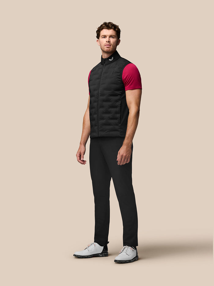 Man wearing black Pinnacle Golf Gilet with stand collar over a magenta tee; sleeveless quilted vest with front zip and Castore branding at the collar and upper back.