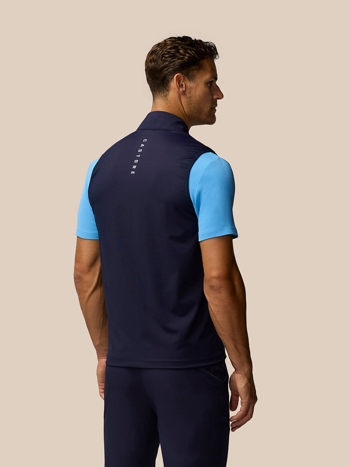 Side view of male model wearing navy Pinnacle Gilet over a light blue polo with back branding on the collar.