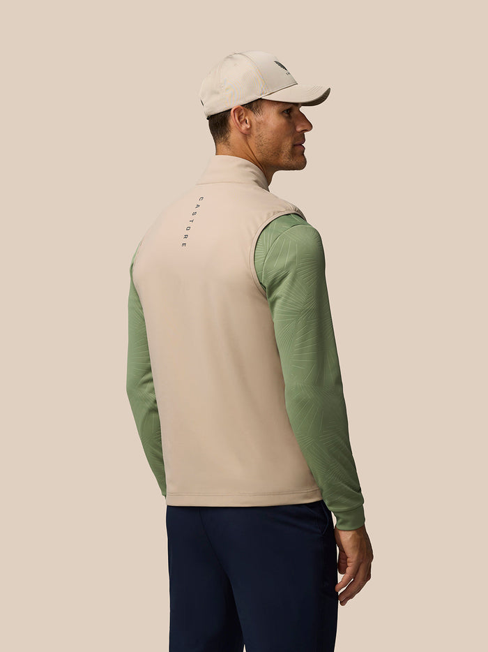 Back view of a man wearing Golf Pinnacle Gilet Mushroom beige vest with green sleeves and a Castore logo on the collar
