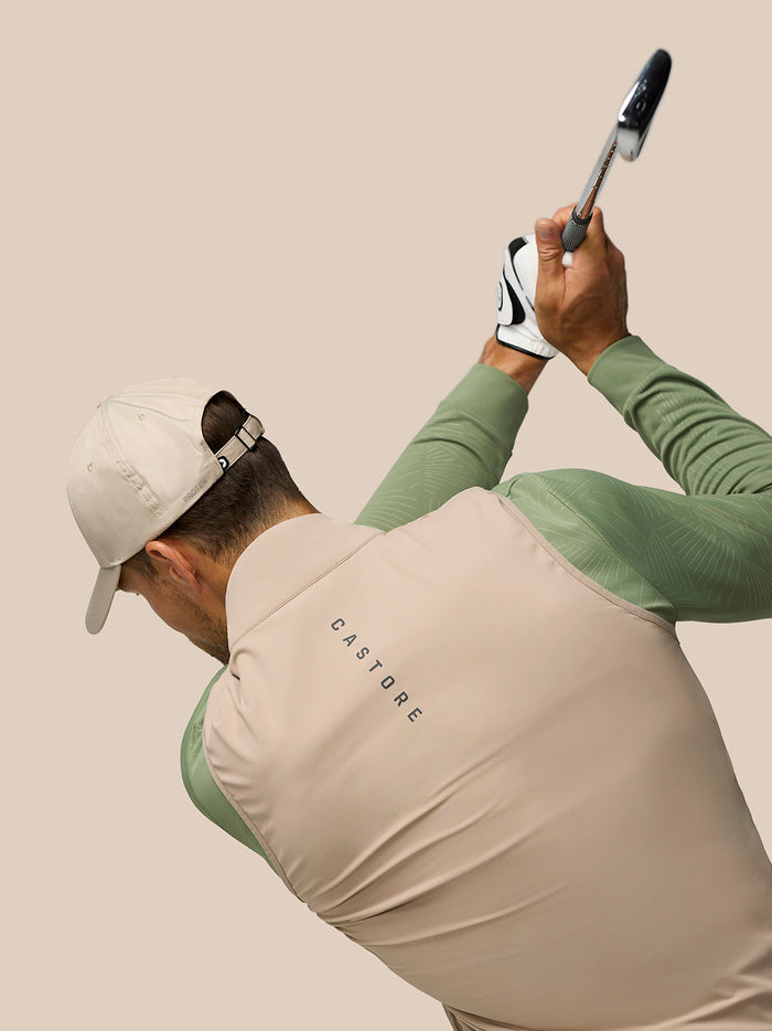 Rear three-quarter view of a golfer wearing Golf Pinnacle Gilet Mushroom beige vest with green sleeves, mid backswing holding a club