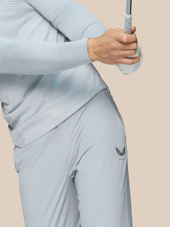 Full-body shot of a man in light grey golf attire wearing Pinnacle trousers and white golf shoes against a beige backdrop.