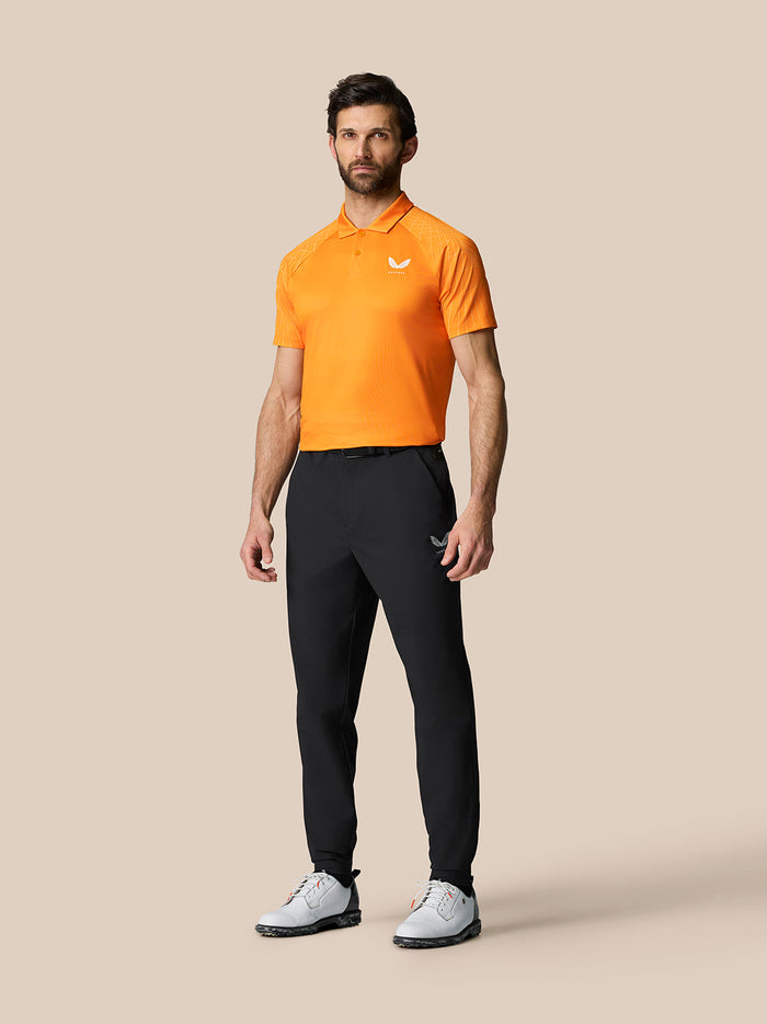 Male model wearing black Golf Pinnacle 32 trousers with an orange polo, Castore branding on the right thigh.