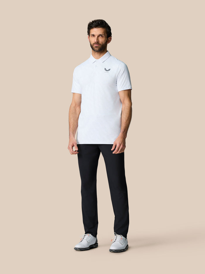 Full-body shot of a man in a white golf ball print Club Graphic Polo with left chest logo and black trousers.