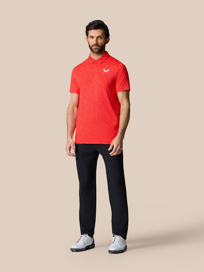 Full body shot of a man wearing Club Graphic Polo in Poppy Red with black trousers on a beige backdrop