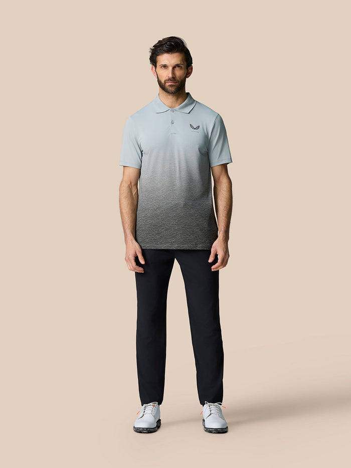 Full front view of a man in a light blue gradient polo with the Castore logo on the left chest, dark trousers and white sneakers.