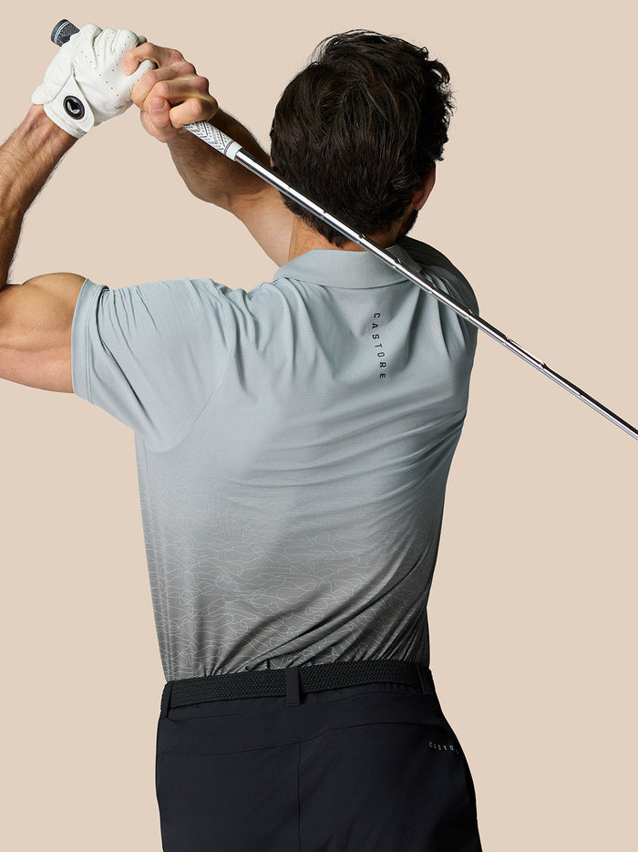 Back view of a man wearing a light grey Castore golf polo with an ombre lower panel and branding along the back, swinging a club.