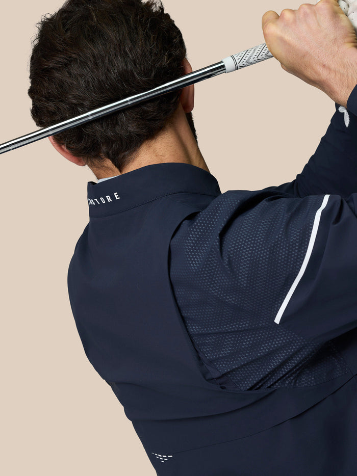 Golfer in navy Golf Pinnacle LVS Woven Jacket swinging a club with back vented sleeves and Castore branding on the collar