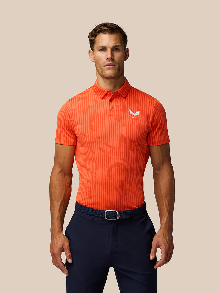 Front view of a man in the bright orange Golf Pinnacle Glow Polo with striped knit and Castore logo on left chest, navy pants.