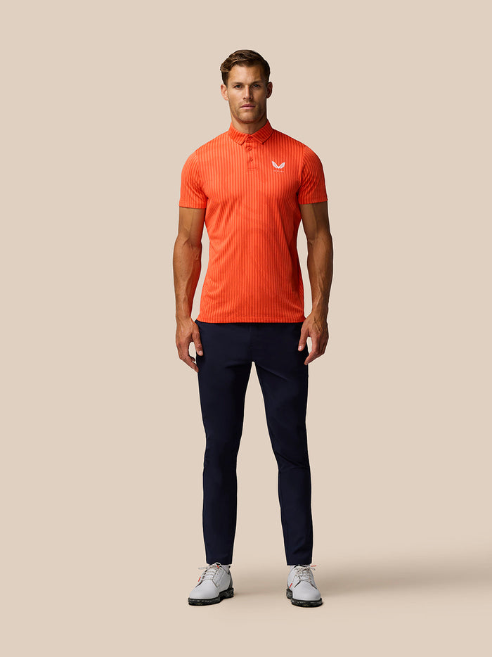 Male model wearing bright orange Golf Pinnacle Glow Polo with vertical stripes and left chest Castore logo.