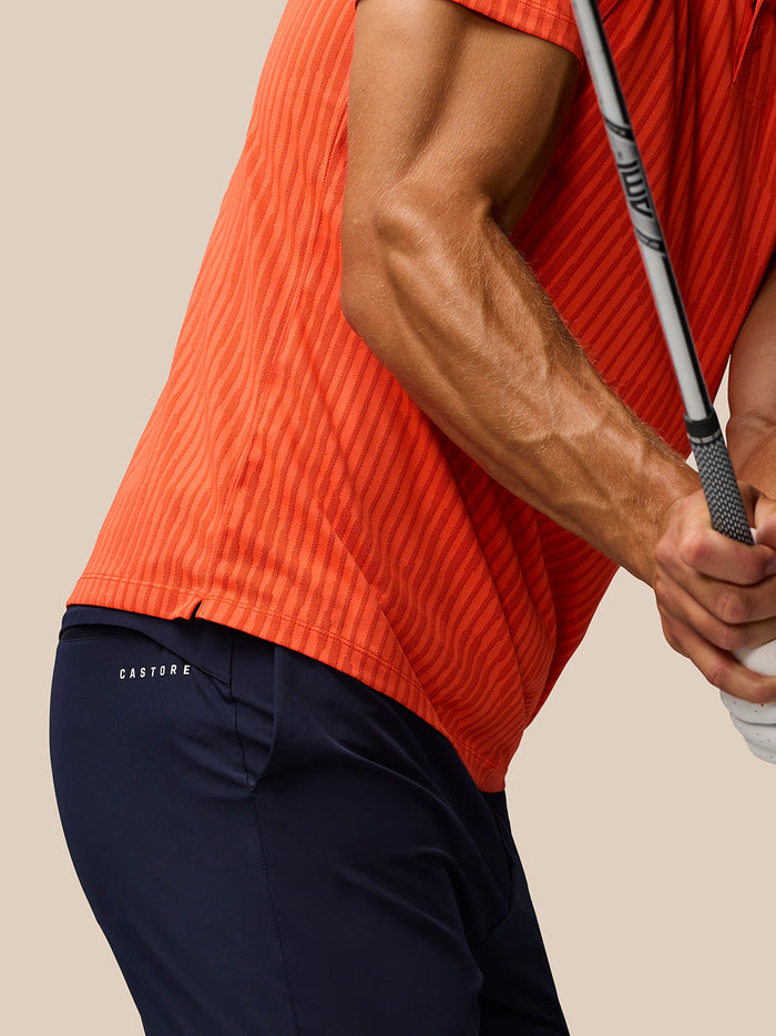 Side view of a man wearing the bright orange Golf Pinnacle Glow Polo with striped knit as he grips a golf club.