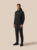 Male model wearing a black golf hoodie with hood and matching pants against a beige background, Castore logo on the chest