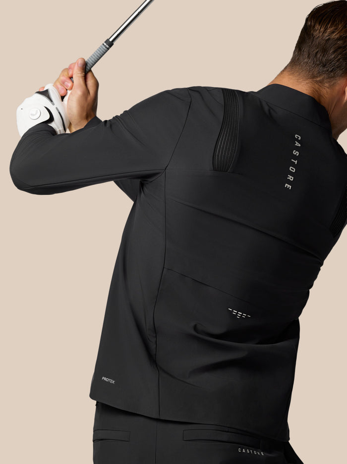 Rear view of a man in a black Castore jacket holding a golf club, showing back vent details and Protek branding.