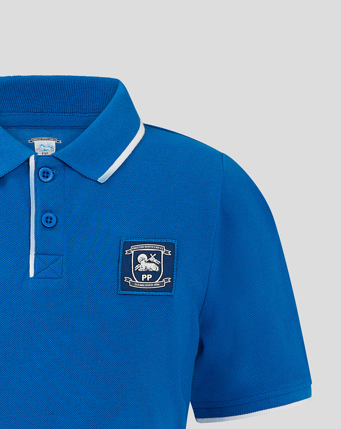 Close-up of blue junior polo showing ribbed collar and crest badge on chest with blue buttons.