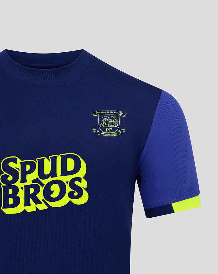 Close-up of the junior away shirt showing the crest on the chest and neon SPUD BROS logo on dark blue fabric.